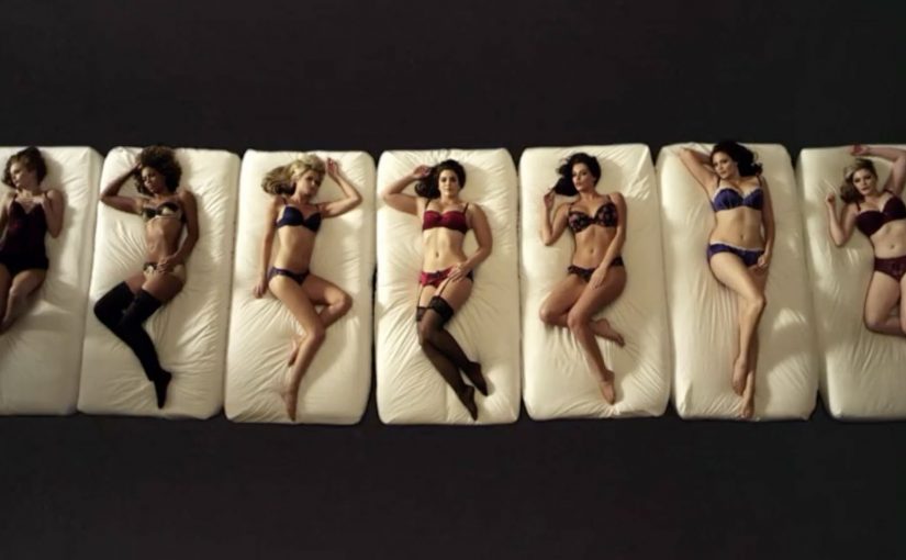 Women in lingerie recline on a piano-like set, singing notes labeled by bra cup size from A to G.