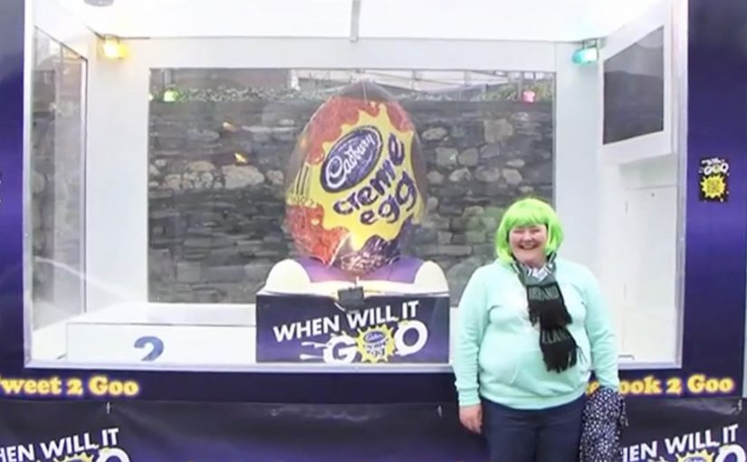 A giant Cadbury Creme Egg in a clear display case on a Dublin street as people gather around it.